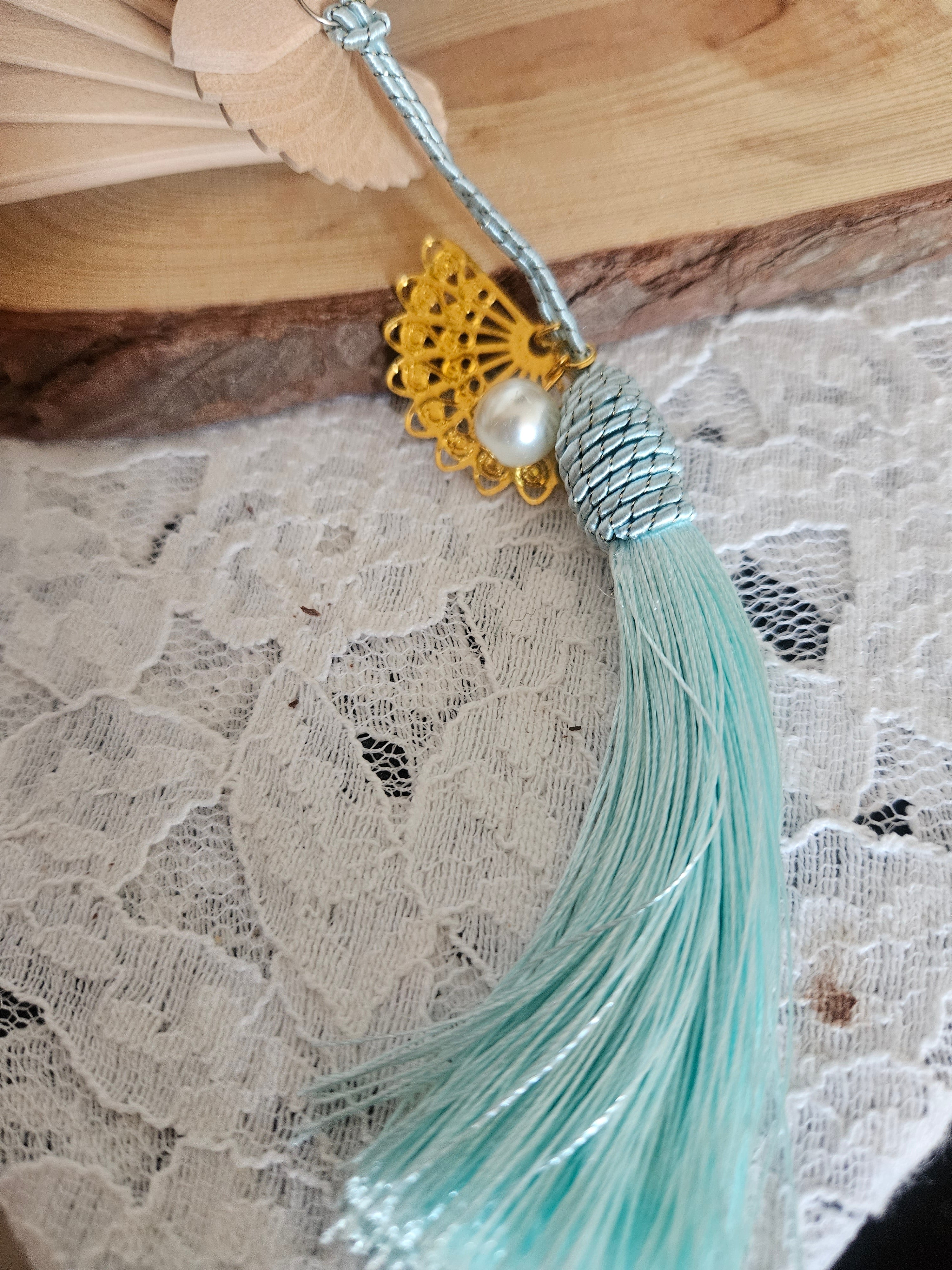 Abanico beige con varilla azul y borla turquesa; detalle dorado con perla en el centro