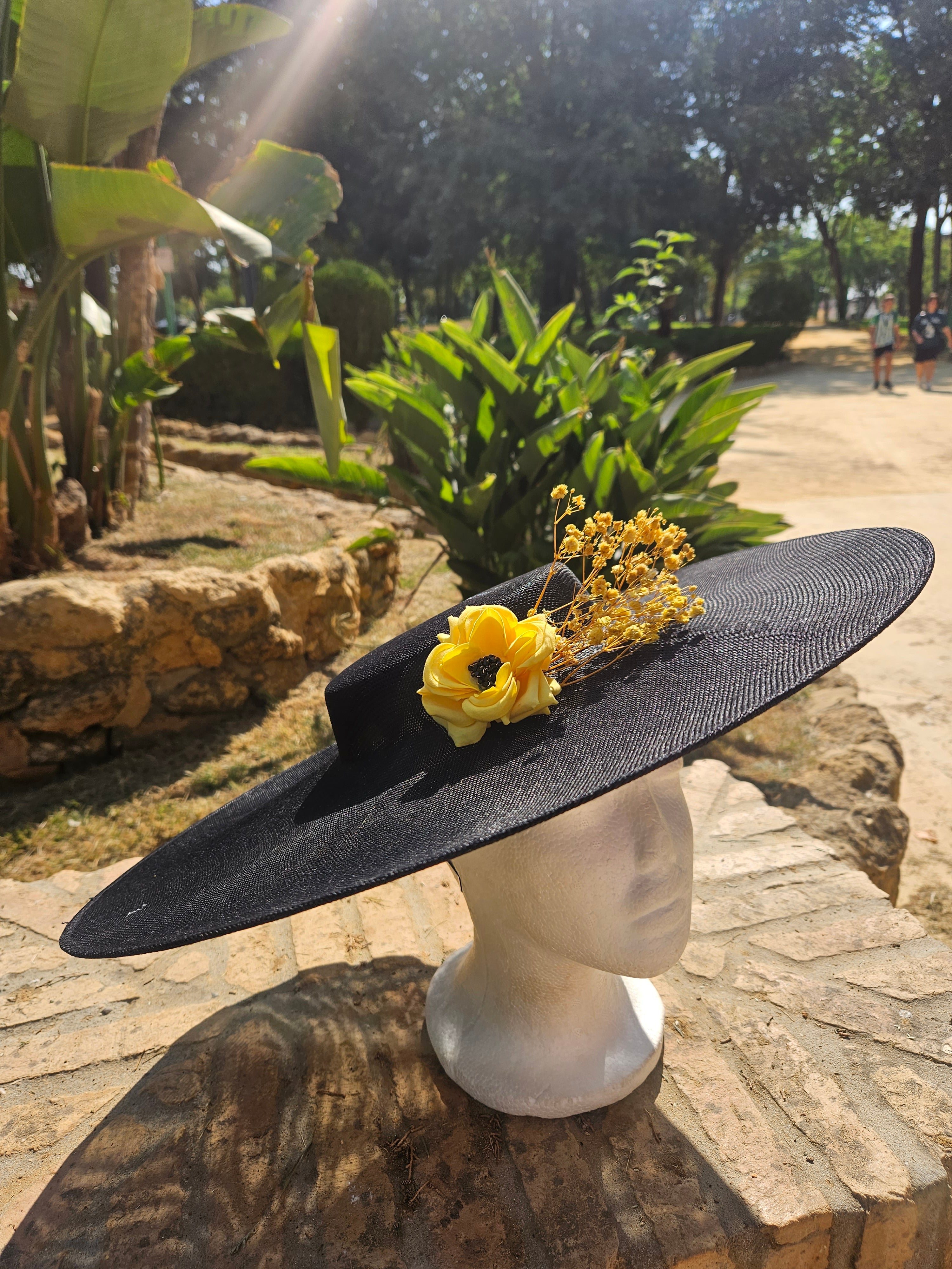 Sombrero de ala ancha negro con flor amarilla y ramitas doradas, vista lateral.