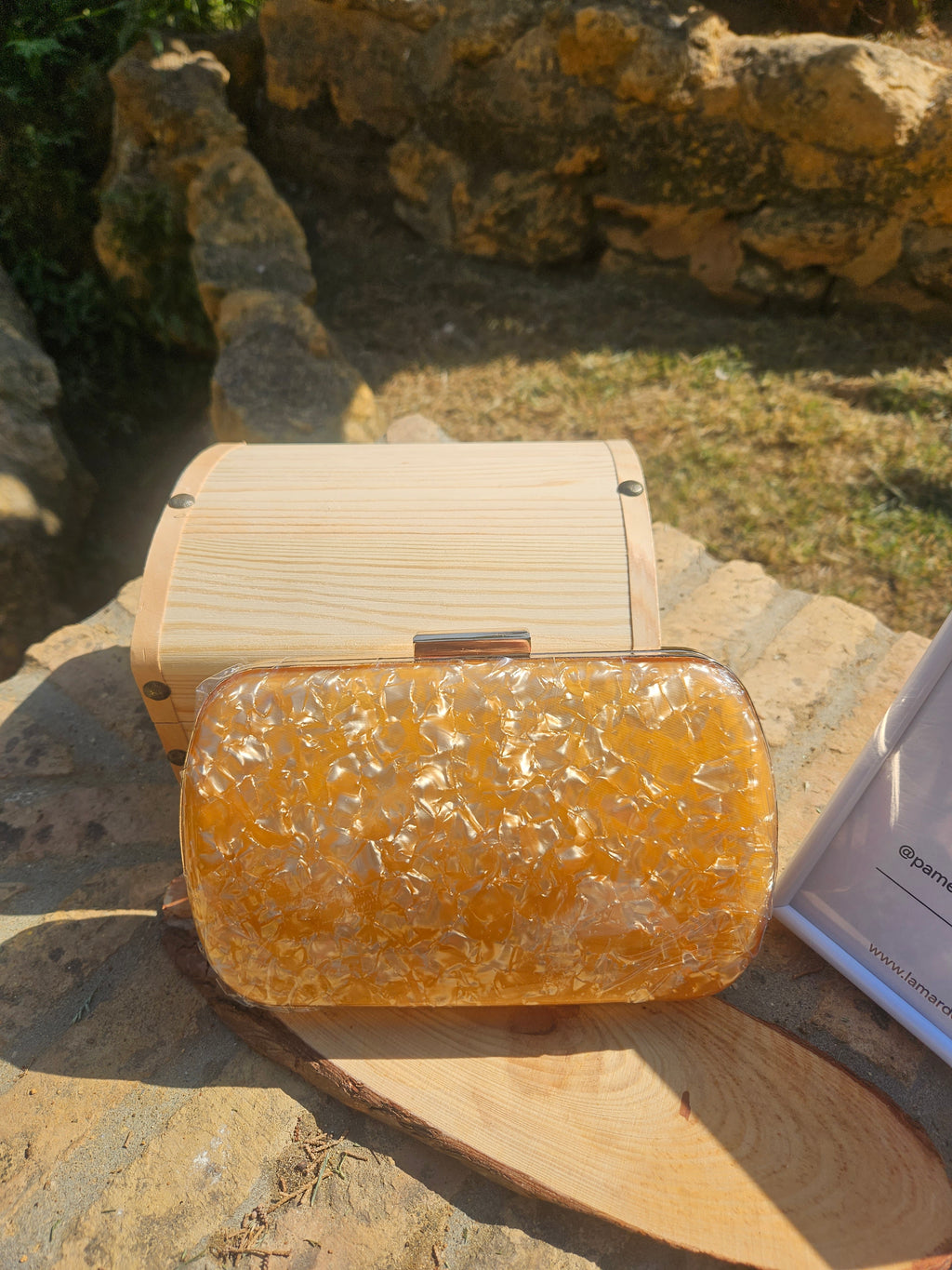Bolso tipo clutch ámbar con cierre metálico y base transparente sobre marco de madera