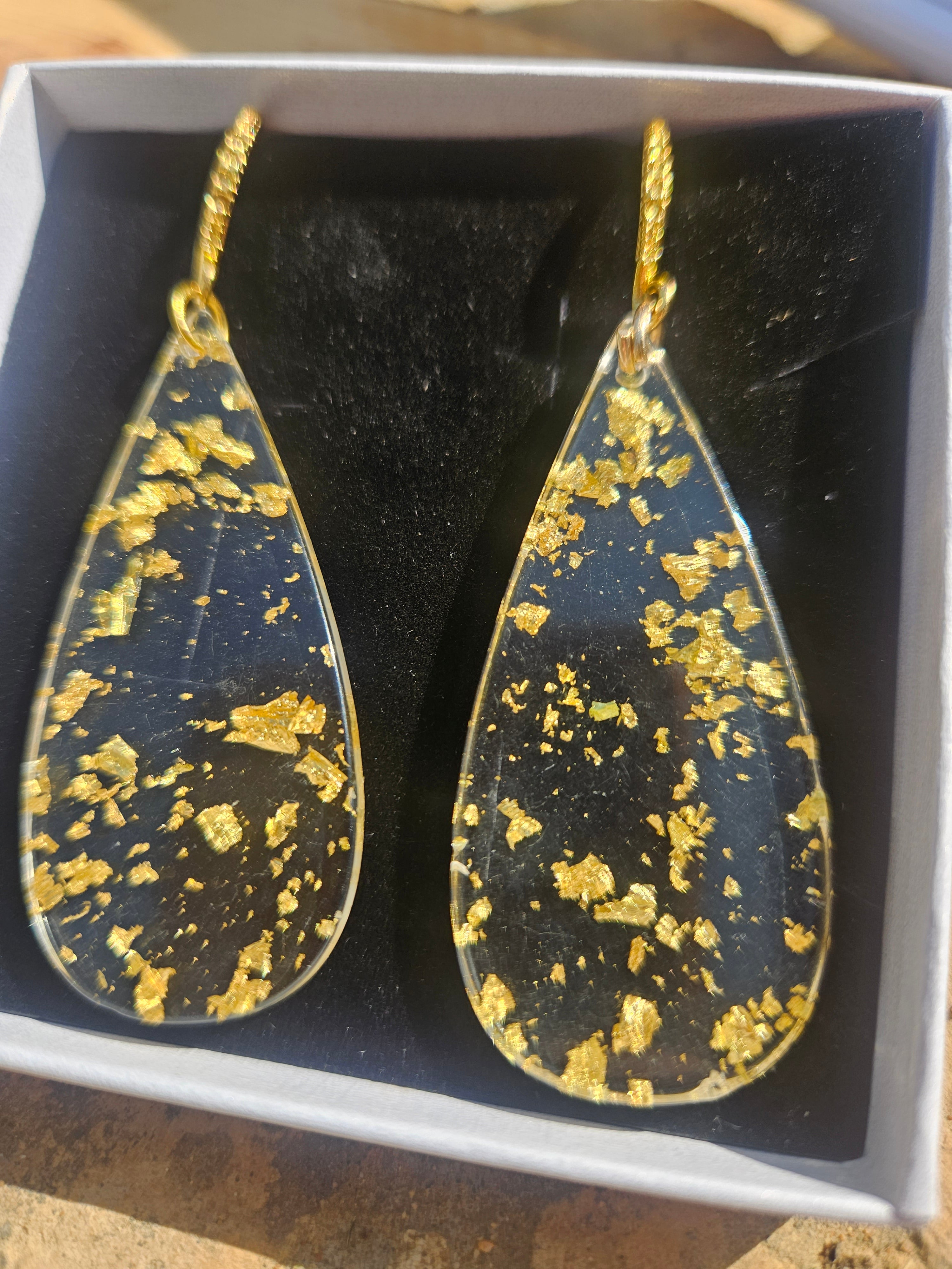 Pendientes de gota negros con salpicaduras doradas y gancho, primer plano en caja.