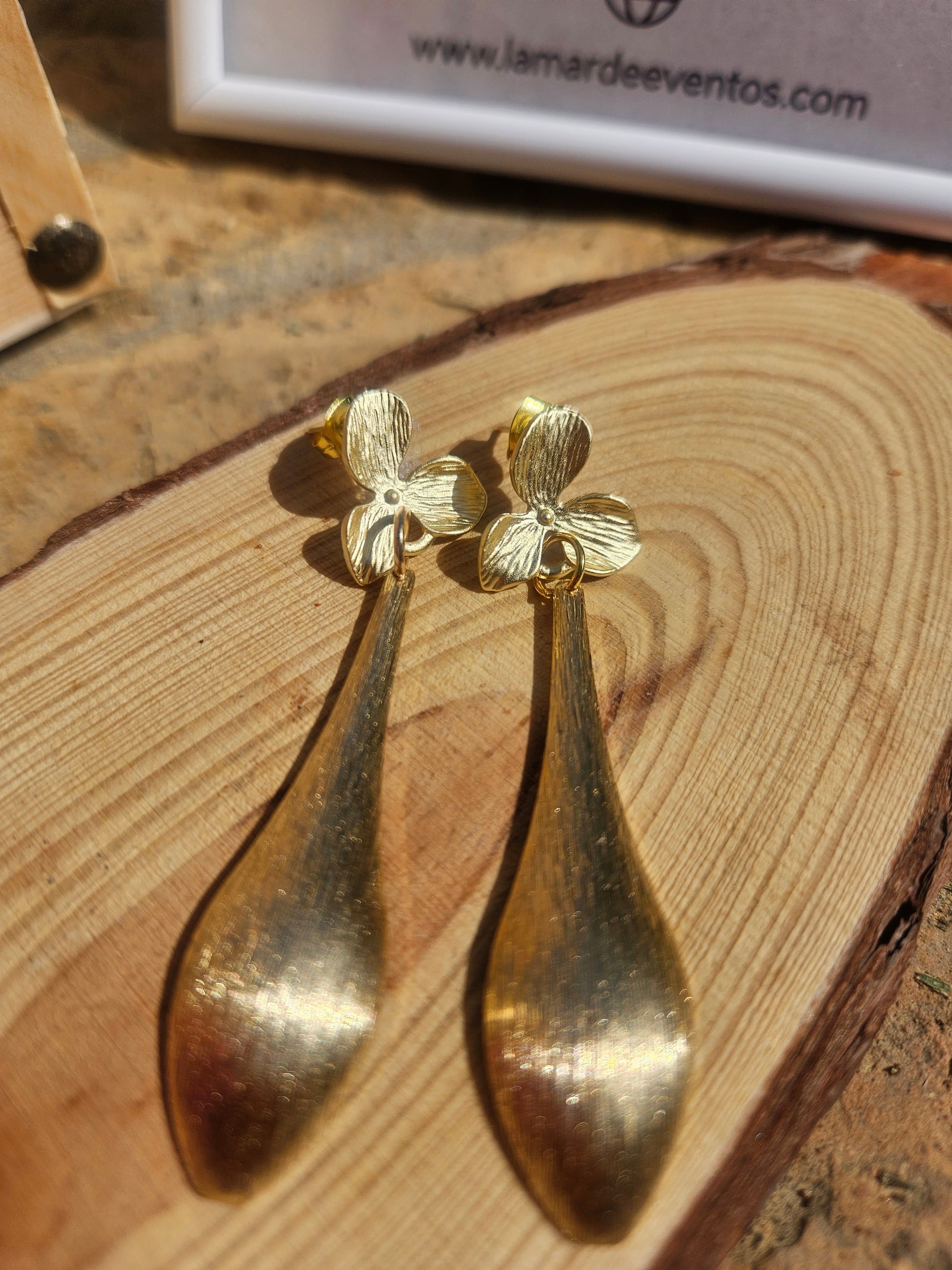 Pendientes dorados con flor y dos colgantes tipo gota, sobre tablón de madera