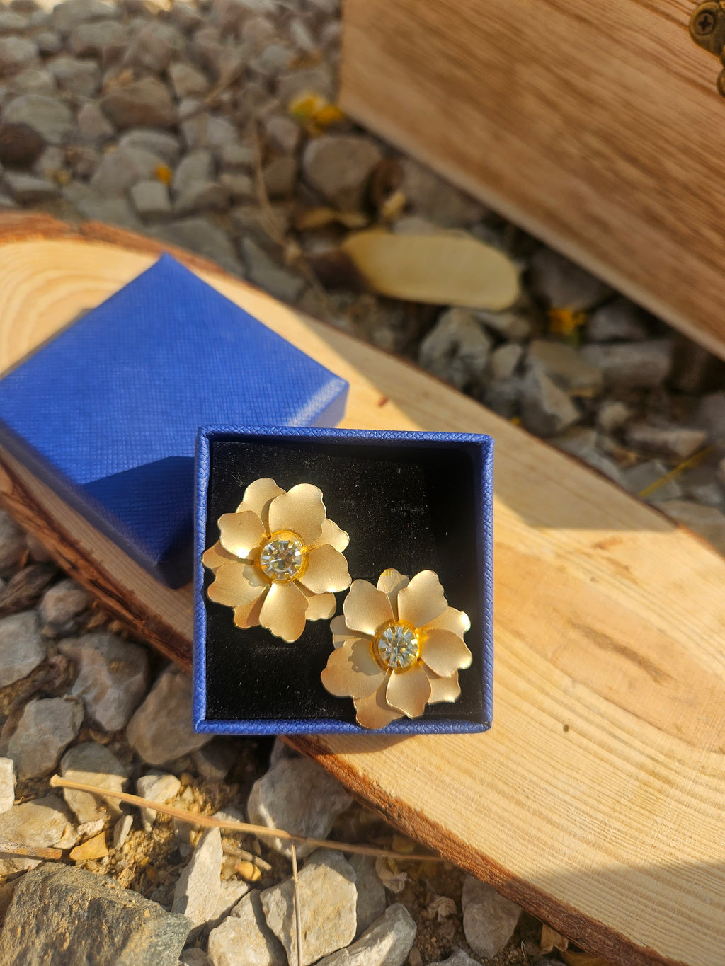 Pendientes con flores doradas y pedrería central, sobre caja negra con tapa azul