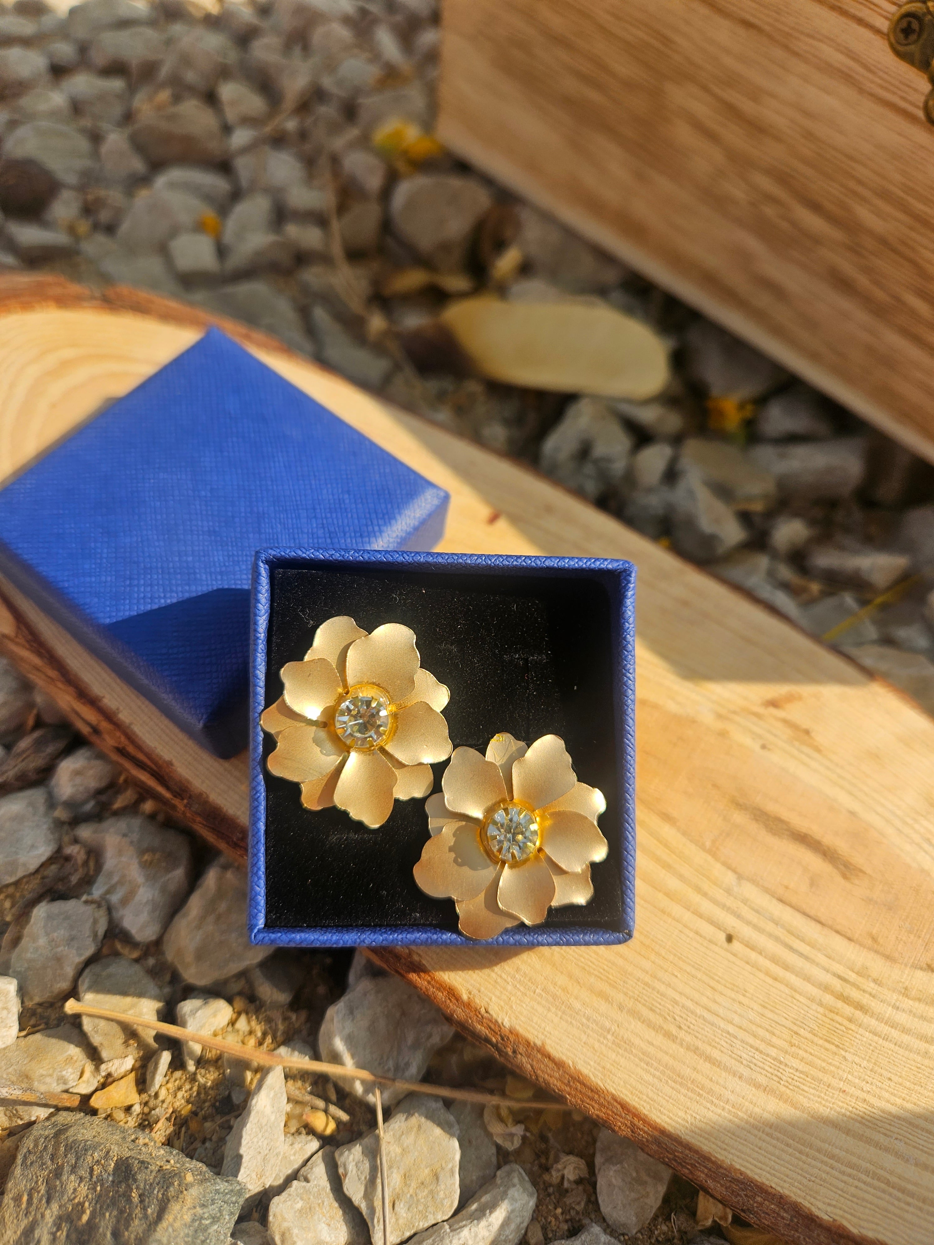 Pendientes con flores doradas y pedrería central, sobre caja negra con tapa azul