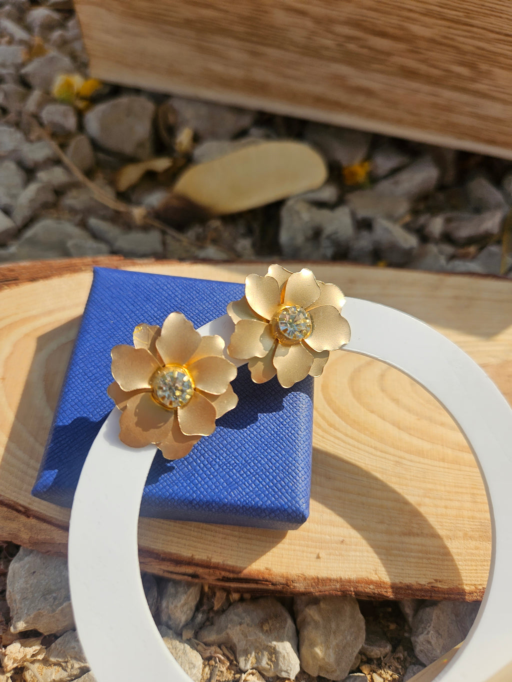 Pendientes con flores doradas y pedrería central, mostrados sobre soporte circular blanco