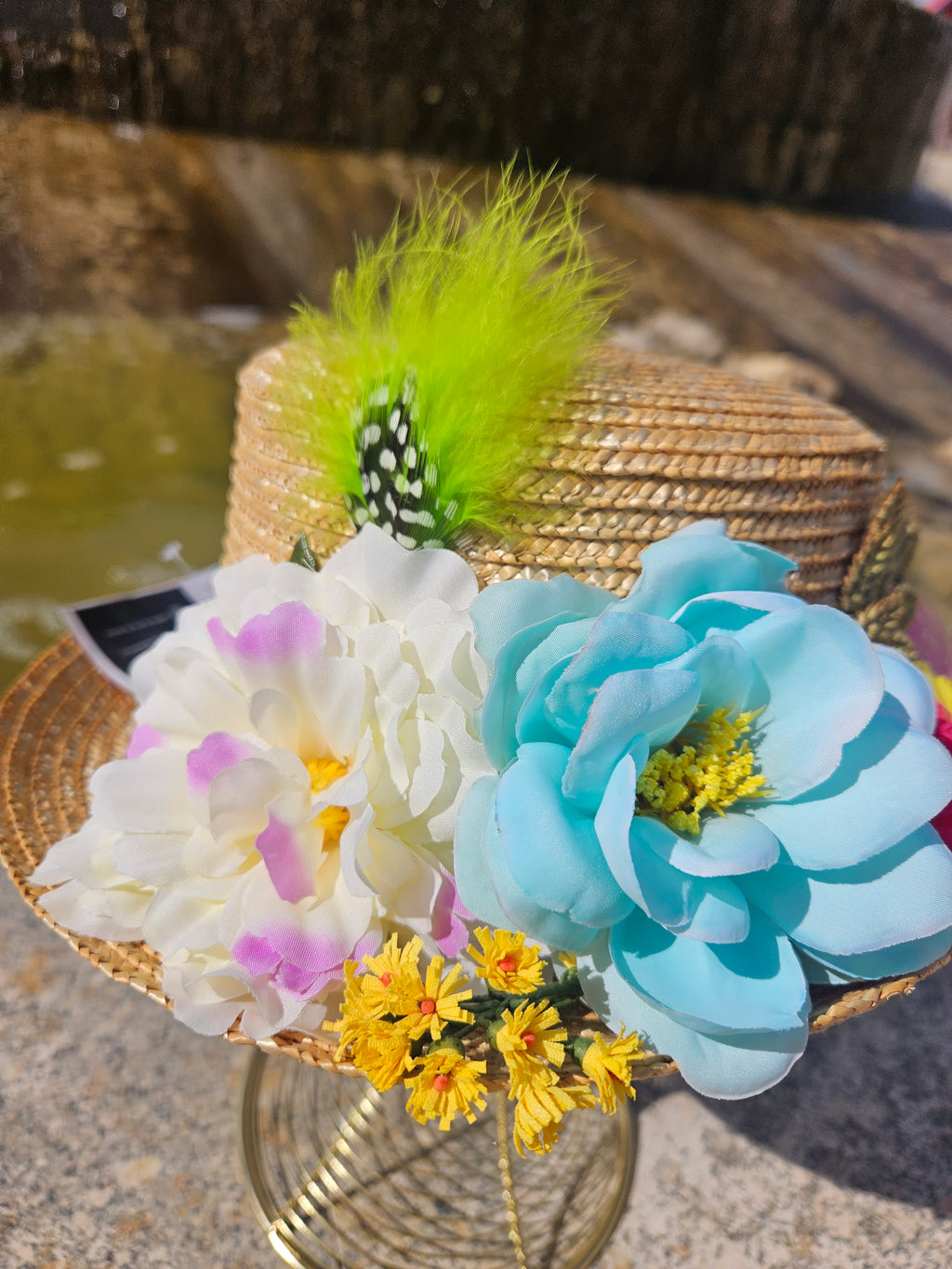 Sombrero de paja beige con flores blancas y azules, margaritas amarillas y pluma verde