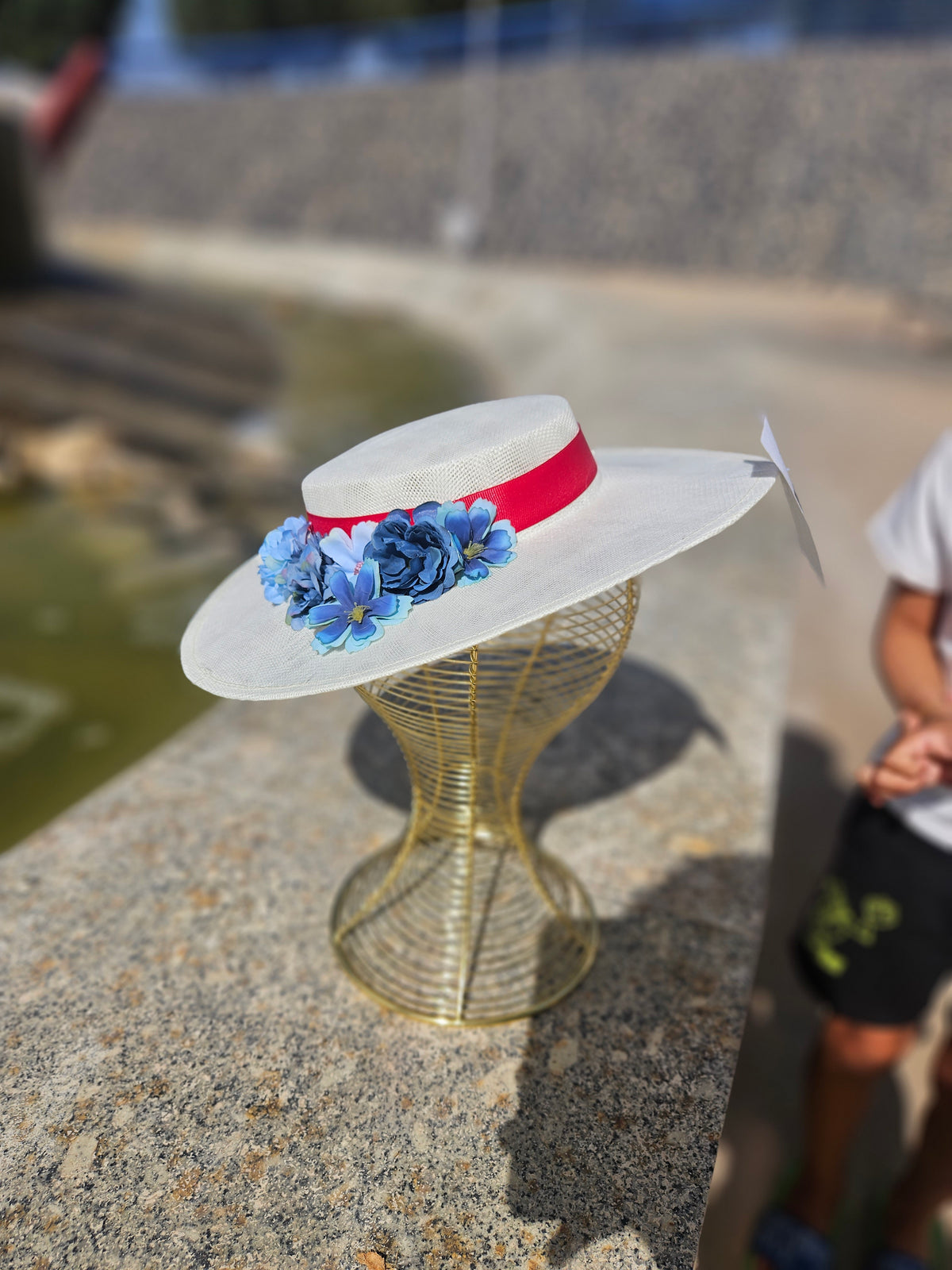 Pamela de ala ancha blanca con flores azules y cinta roja, puesta sobre maniquí