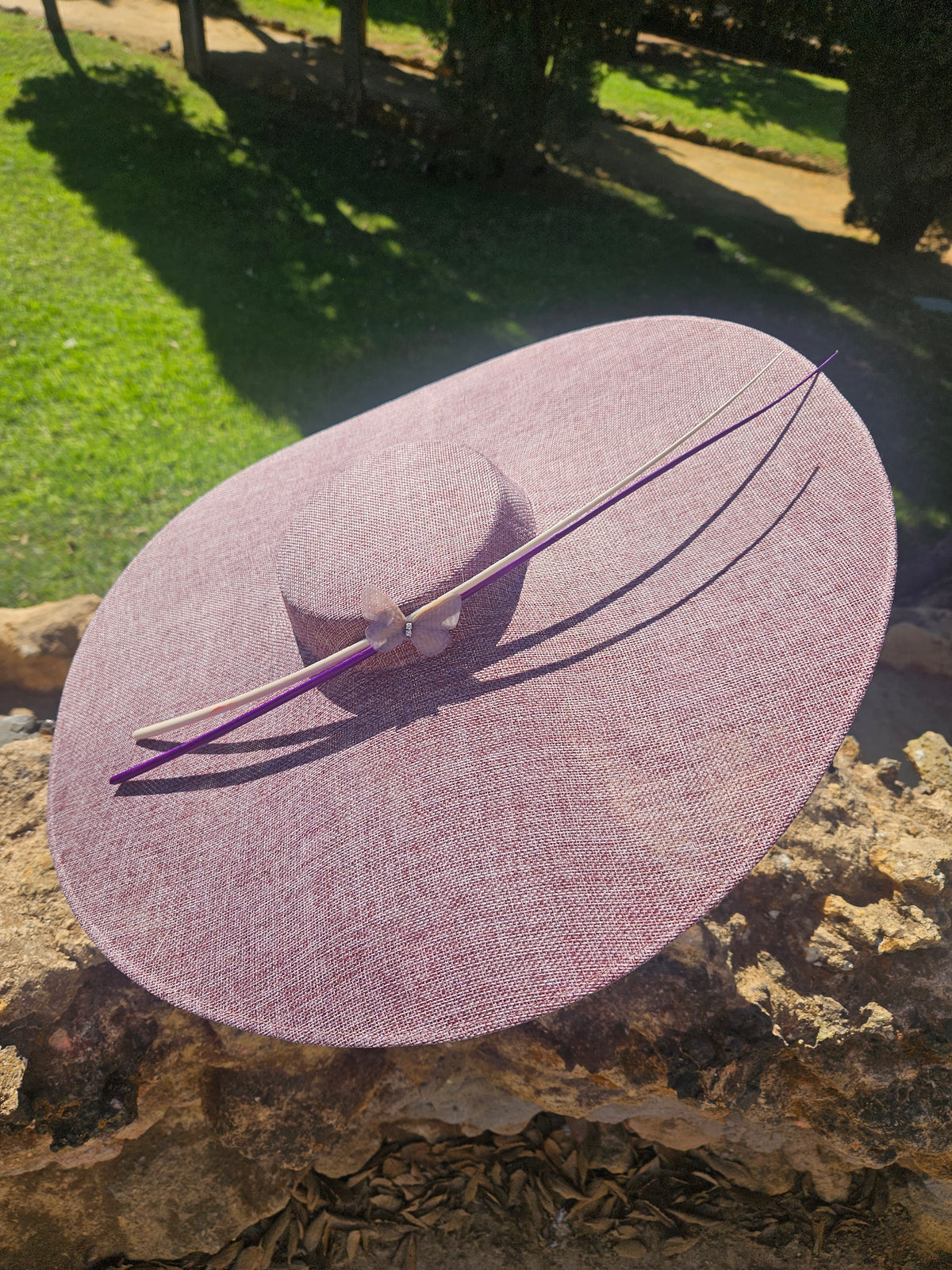 Sombrero pamela rosa de ala ancha con cinta morada y adorno floral en la copa.