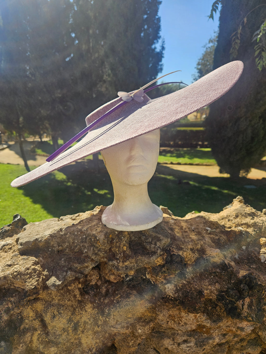 Sombrero pamela rosa claro de ala ancha con cinta morada y lazo, sobre maniquí.