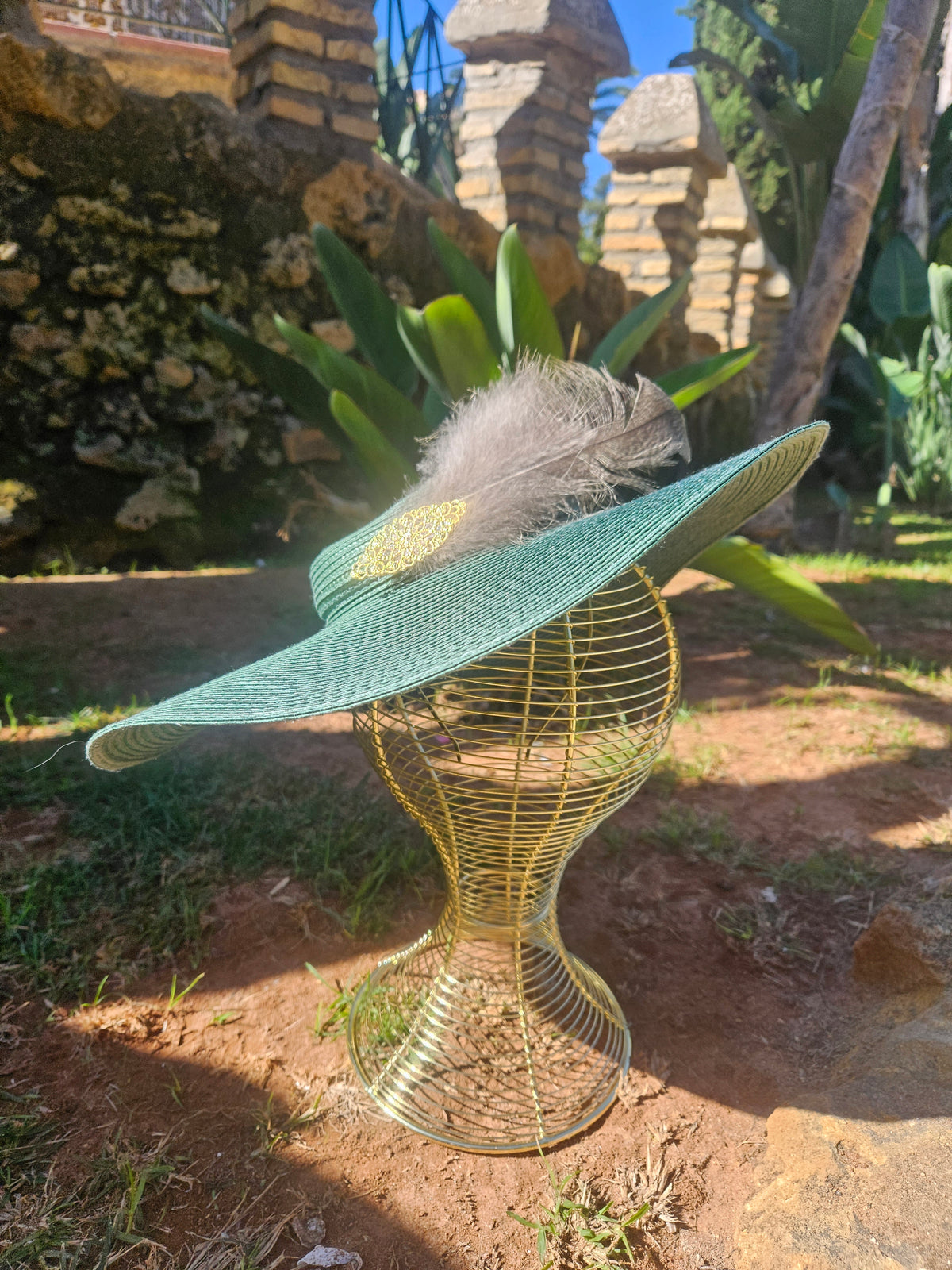 Sombrero turquesa de ala ancha con pluma gris y adorno dorado sobre maniquí